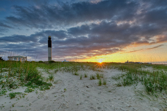 Oak Island Lighthouse at Caswell Beach, North Carolina, under a cloudy sunrise sky with sunlight illuminating the scene.